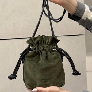 May include: An olive green suede drawstring bucket bag with a black leather shoulder strap and matching black leather drawstring cords. The bag features a gathered top closure and the embossed text SQUAMPLI on the lower front section.