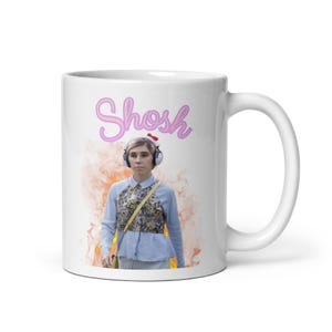 May include: White ceramic mug with a person wearing headphones and a floral top. The word "Shosh" is written in pink cursive above the person. The mug has a white handle and a colorful background.