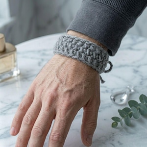 May include: A gray crochet bracelet is worn on a wrist. The bracelet is made of a thick, textured yarn and has a simple tie closure. The bracelet is a neutral color and appears handmade.