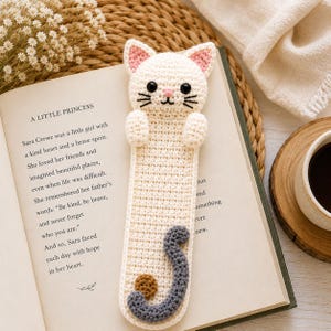 May include: A cream-colored crochet bookmark shaped like a cat with a white head, pink inner ears, black embroidered whiskers, and dark eyes. The body is a long, flat strip featuring a gray crocheted tail and a small brown yarn ball at the bottom.