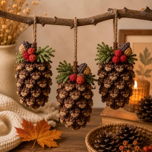 Könnte beinhalten: Drei gehäkelte Tannenzapfen-Ornamente, die an einem Holzast hängen. Jeder braun-beige Zapfen ist mit grünen gehäkelten Nadeln, kleinen roten Beeren und einer kleinen Eichelkappe verziert und an einer rustikalen Juteschnur aufgehängt.