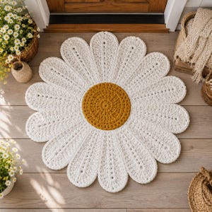 May include: A round, crocheted rug shaped like a daisy flower, featuring a textured mustard yellow center surrounded by numerous white, elongated petals with visible stitch detailing throughout the entire floral design.