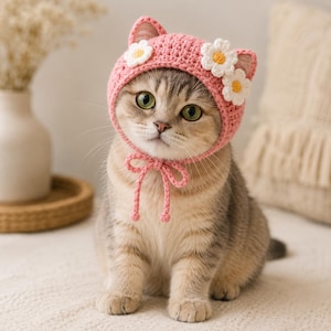 May include: A pink crocheted cat-ear bonnet featuring two white daisy appliques with yellow centers on the side. The bonnet has integrated ear-shaped points and a matching pink crocheted chin strap that ties into a bow under the chin.