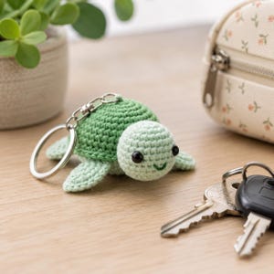 May include: A small, crocheted turtle keychain featuring a dark green shell and a light green head with black plastic eyes and a stitched green smile. The turtle is attached to a silver-colored metal split ring and a lobster claw clasp.