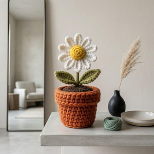 Puede incluir: Una flor de margarita de ganchillo hecha a mano en una maceta naranja. La margarita tiene pétalos blancos y un centro amarillo. La maceta está llena de hilo marrón para simular tierra, y la flor tiene hojas verdes. La flor de ganchillo es un artículo decorativo.