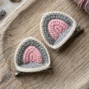 May include: Two hair clips featuring a triangular crochet design with a light pink centre, a dark grey border, and an off-white outer edge. Each piece is attached to a silver-toned metal alligator clip, showcasing a layered, textured yarn construction.