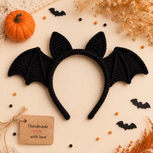 May include: A black crochet headband featuring two pointed bat ears and two large, textured bat wings extending from the sides. A rectangular brown paper tag is attached, printed with the text Handmade with love and a small heart icon.