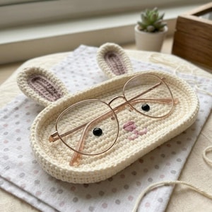 May include: An oval-shaped, cream-colored crochet tray featuring two upright bunny ears, two small black bead eyes, and a pink stitched nose. A pair of thin, gold-rimmed round eyeglasses rests on top of the tray, positioned over the facial features.
