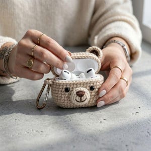 May include: A beige crochet bear-shaped case for wireless earbuds. The case is open, revealing white earbuds inside. The case has a brown nose and black eyes. A gold-colored clasp is attached to the case.