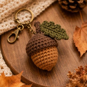May include: A crochet acorn keychain featuring a light brown nut, a dark brown cap, and a textured dark green leaf. The acorn is attached to a bronze-toned metal split ring and a lobster clasp, providing a secure way to attach the item to keys.