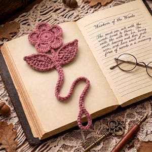 Könnte beinhalten: Ein altrosa gehäkeltes Blumen-Lesezeichen mit langem Stiel und einer Schlaufe am Ende, das auf einem offenen Buch mit handschriftlichem Text mit dem Titel Wisdom of the Woods liegt, begleitet von einer runden Brille und einem Metallschlüssel.