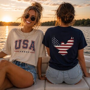 May include: Two women wearing patriotic t-shirts on a boat at sunset. One shirt is beige with "USA" in red and blue lettering. The other is navy blue with an American flag eagle design on the back. Both are wearing denim shorts.