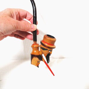 May include: A wooden tobacco pipe with a carved face design. The pipe features a black stem and a bowl shaped like a man's face wearing a hat with a red band. The pipe has a red accent and is made of light and dark wood.