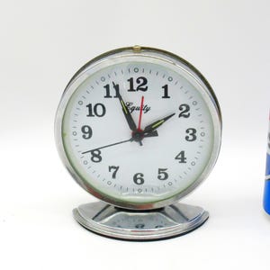 May include: A round analog alarm clock with a silver-tone metal frame and a white face featuring black Arabic numerals. The clock has black hour and minute hands, a red second hand, and the word Equity printed on the dial. A 12-ounce soda can is shown.