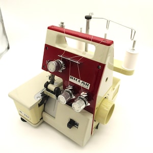 May include: A cream and burgundy Riccar Lock BL-233 serger sewing machine. The machine has multiple dials and a handle. The text "Riccar Lock" is visible on the front. Two spools of white thread are attached.