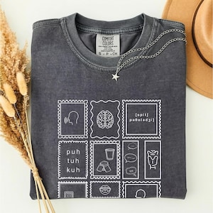 May include: Dark grey Comfort Colors t-shirt with a white graphic of postage stamp-like designs. The shirt has a crew neck and short sleeves. A silver necklace with a star pendant and a brown hat are also visible.