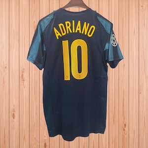 May include: A dark blue sports jersey featuring the name ADRIANO and the number 10 in yellow lettering on the back. The shirt has dark teal side panels and a circular patch on the right sleeve. The design includes a perforated texture on the numbers.