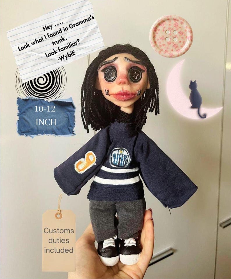 May include: A doll with black yarn hair, button eyes, and stitched mouth details, wearing a navy blue long-sleeve shirt with white stripes and a logo, grey pants, and black and white shoes. The doll measures 10-12 inches in height.