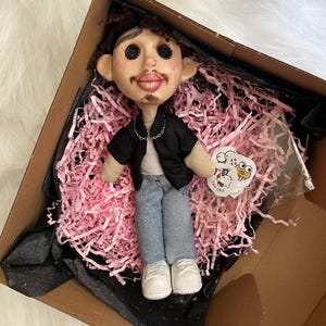 May include: A handmade doll with brown curly hair, button eyes, and a mustache, wearing a black open shirt over a white top, blue jeans, and white shoes. The doll features a silver chain necklace and is presented in a box with pink paper filler.