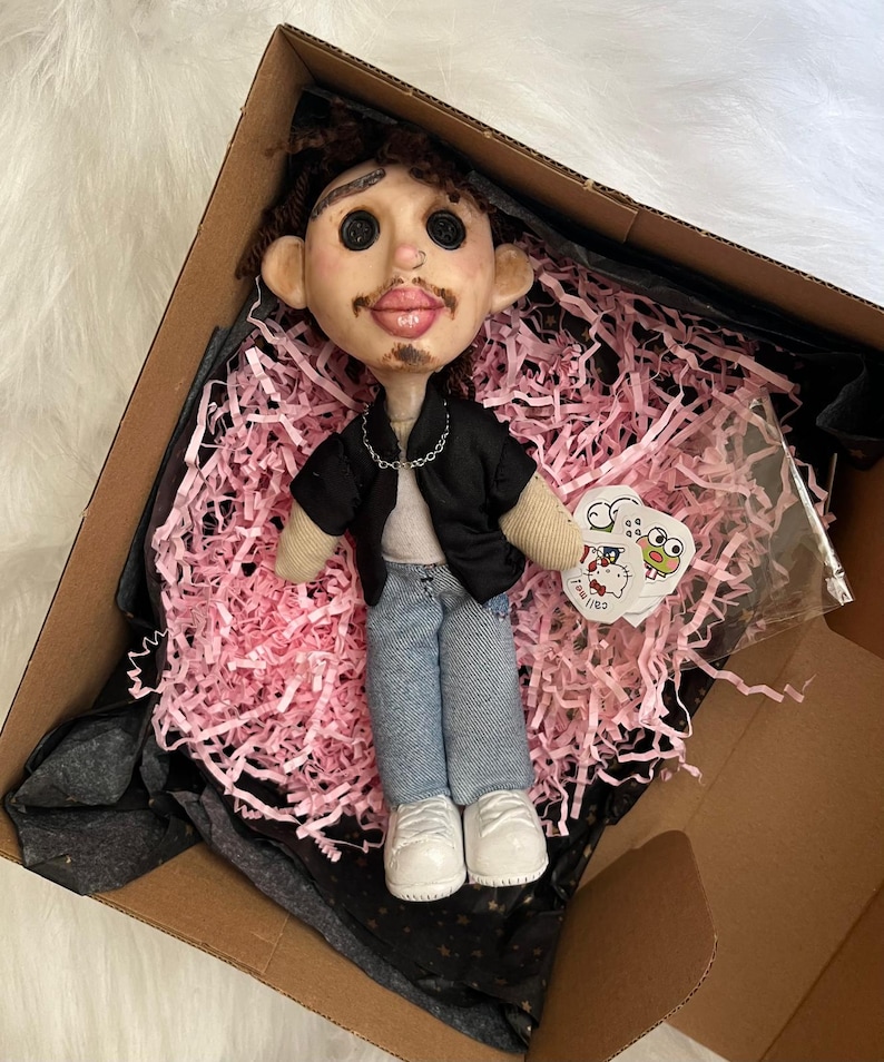 May include: A handmade doll with brown curly hair, button eyes, and a mustache, wearing a black open shirt over a white top, blue jeans, and white shoes. The doll features a silver chain necklace and is presented in a box with pink paper filler.