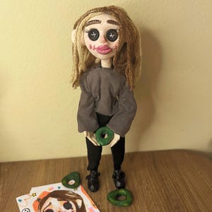 May include: A handmade doll with light skin, long brown yarn hair, and black button eyes. The doll wears a gray long-sleeved shirt, black pants, and black shoes. It holds a small green ring-shaped object, with two additional green rings placed near its feet.
