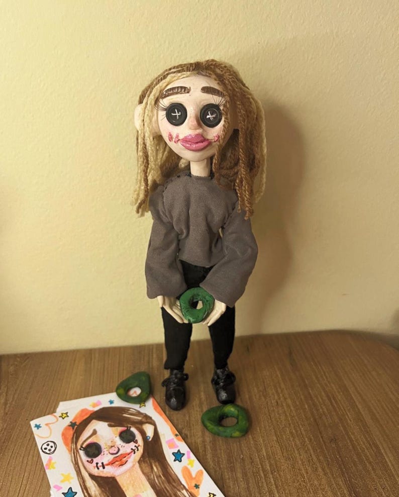 May include: A handmade doll with light skin, long brown yarn hair, and black button eyes. The doll wears a gray long-sleeved shirt, black pants, and black shoes. It holds a small green ring-shaped object, with two additional green rings placed near its feet.