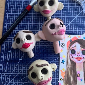May include: Four sculpted clay doll heads with button eyes, painted eyebrows, and pink lips, alongside a paintbrush and a colorful illustrated portrait. The heads feature various facial details including small metal piercings and stitched mouth designs.