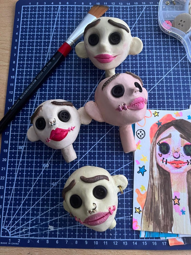May include: Four sculpted clay doll heads with button eyes, painted eyebrows, and pink lips, alongside a paintbrush and a colorful illustrated portrait. The heads feature various facial details including small metal piercings and stitched mouth designs.