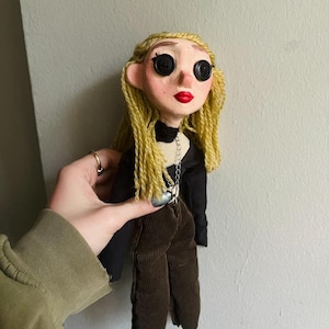 May include: A handmade doll with pale skin, bright red lips, and black button eyes. The doll has long, wavy blonde yarn hair and wears a black long-sleeved top with a silver chain necklace and dark brown corduroy pants with a textured ribbed finish.