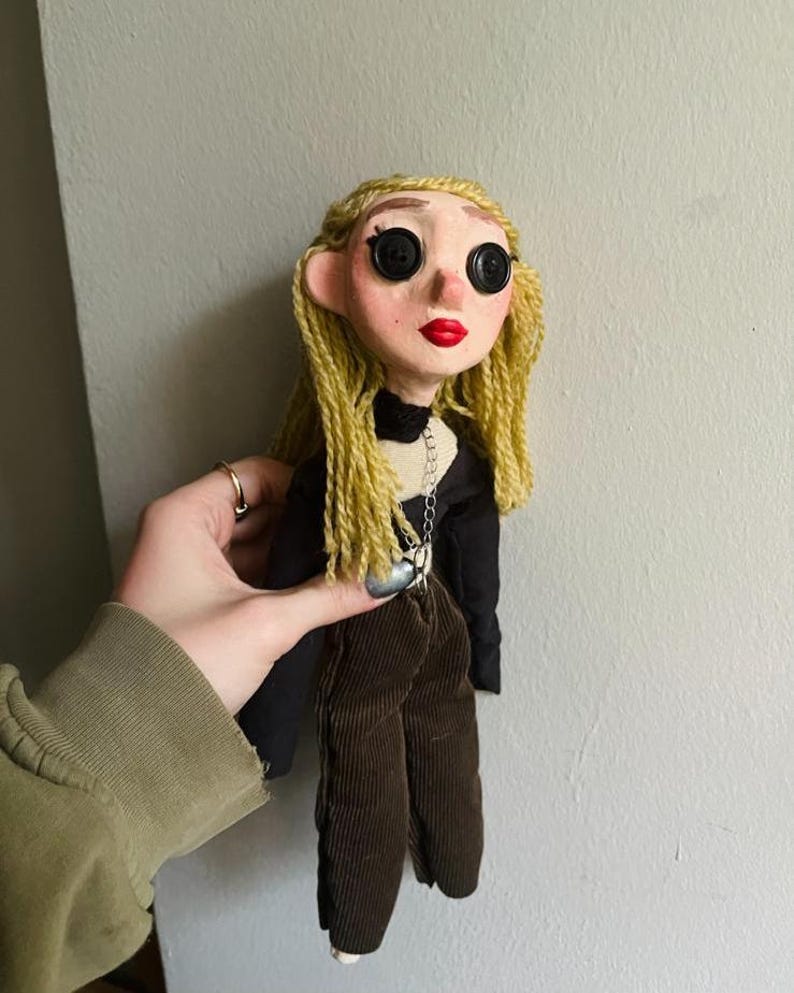 May include: A handmade doll with pale skin, bright red lips, and black button eyes. The doll has long, wavy blonde yarn hair and wears a black long-sleeved top with a silver chain necklace and dark brown corduroy pants with a textured ribbed finish.
