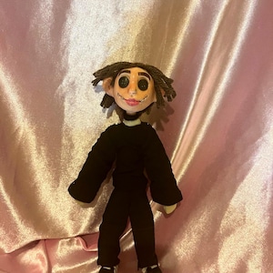 May include: A handmade doll with dark brown yarn hair, large black button eyes, and a stitched mouth. The doll wears a long-sleeved black shirt, black trousers, and black and white sneakers. The facial features are sculpted with a light skin tone.