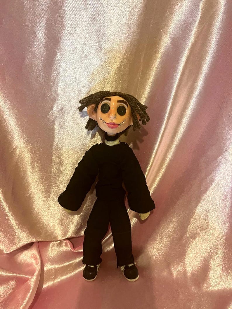 May include: A handmade doll with dark brown yarn hair, large black button eyes, and a stitched mouth. The doll wears a long-sleeved black shirt, black trousers, and black and white sneakers. The facial features are sculpted with a light skin tone.