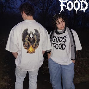 Puede incluir: Dos personas con camisetas blancas con diseños gráficos. Una camiseta presenta una figura alada sobre una hoguera, la otra muestra el texto "GODS FOOD" en una fuente gótica. Ambos llevan vaqueros de color claro.