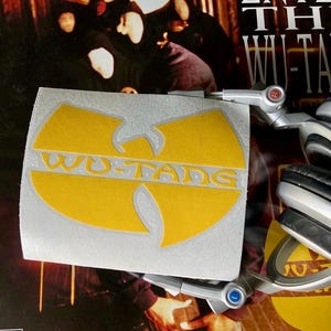 May include: A yellow Wu-Tang Clan logo sticker with the text "WU-TANG" in a bold font. The sticker is on a silver background, with a record album and headphones in the background.