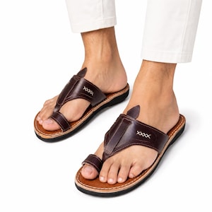 May include: Dark brown leather sandals featuring a toe loop design, a wide strap across the top of the foot, and decorative white cross-stitch detailing. The sandals have a tan leather footbed with visible stitching along the edge and a black sole.