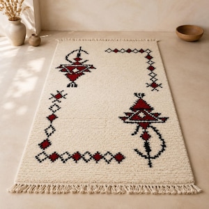 May include: A rectangular off-white area rug featuring a textured pile and a decorative border pattern in deep red and black. The design includes geometric shapes and stylized motifs, finished with knotted fringe along the short edges.