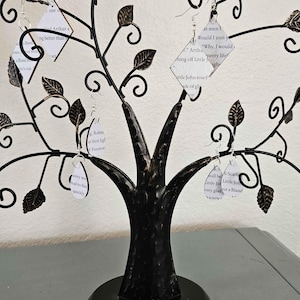 May include: Black metal earring display in the shape of a tree, with leaf-shaped accents. The tree holds several pairs of earrings, each with a small tag featuring printed text. The base is round.
