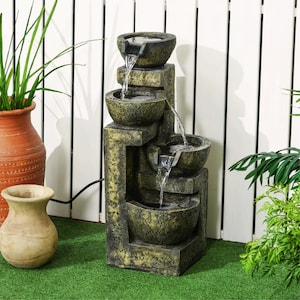 Könnte beinhalten: Ein mehrstufiger Outdoor-Wasserbrunnen mit verwitterter Steinoptik. Wasser fließt von der oberen Schale durch drei untere Schalen. Der Brunnen steht auf einer grünen Oberfläche mit einem weißen Zaun im Hintergrund. Eine Terrakotta-Vase und eine kleine beige Vase befinden sich links.