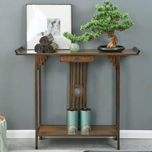 Może przedstawiać: Drewniana konsola z bonsai w czarnej doniczce, oprawionym zdjęciem i zwiniętymi matami. Stół posiada szufladę i dolną półkę z dwoma turkusowymi pojemnikami. Stół wykonany jest z ciemnego drewna.