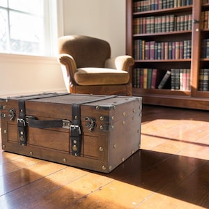 May include: A rectangular wooden storage chest featuring a dark brown finish, reinforced metal corner accents, and two black leather-like straps with silver-toned buckles. The chest includes a central handle and metallic latches on the front panel.