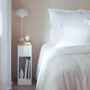 May include: A white bedside table with a wooden top, holding a lit candle in a glass holder, a white vase, and books. A white lamp with a unique shade is next to the table. The bed has white bedding.