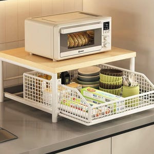 Puede incluir: Estantería de cocina blanca y de madera con un pequeño horno encima. La estantería tiene una cesta de alambre extraíble llena de utensilios de cocina, cuencos y utensilios. La puerta del horno está abierta, revelando productos horneados.