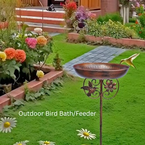 May include: A bronze-effect bird bath with a hummingbird and flower design. The bird bath is on a tall stand, set in a garden with green grass and flowers. The text "Outdoor Bird Bath/Feeder" is visible.
