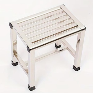 May include: A stainless steel step stool with a rectangular top and four legs. The top has horizontal slats, and the legs have black rubber feet. The stool is designed for stability and support.