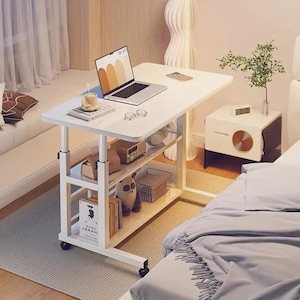 Könnte beinhalten: Ein weißer, rollbarer Laptop-Schreibtisch mit drei Regalen, auf denen Bücher, ein Stofftier und andere Gegenstände stehen. Ein Laptop, eine Kaffeetasse und eine Brille stehen auf der Oberfläche des Schreibtischs. Der Schreibtisch befindet sich in einem Schlafzimmer.
