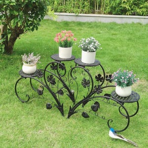 Peut inclure: Support de plantes en fer forgé noir avec cinq plateformes circulaires, chacune contenant une plante en pot avec des fleurs colorées. Le support présente des motifs décoratifs de feuilles et de vignes, sur une pelouse.