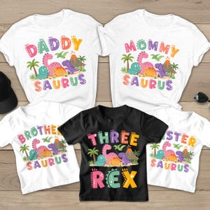 May include: A collection of five t-shirts featuring colorful dinosaur graphics, palm trees, and volcanoes. The shirts display text including DADDY SAURUS, MOMMY SAURUS, BROTHER SAURUS, SISTER SAURUS, and THREE REX in vibrant, multi-colored block lettering.
