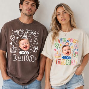 May include: A dark brown t-shirt with white text reading IT'S FUN TO BE DADA and a cream t-shirt with colorful text reading IT'S FUN TO BE MAMA. Both shirts feature a graphic of a baby wearing a party hat, a cake, balloons, and a gift box.
