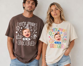 以下が含まれることがあります： IT'S FUN TO BE DADAと白文字で書かれたダークブラウンのTシャツと、IT'S FUN TO BE MAMAとカラフルな文字で書かれたクリーム色のTシャツ。どちらのTシャツにも、パーティーハットをかぶった赤ちゃんのグラフィック、ケーキ、風船、ギフトボックスが描かれています。