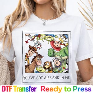 May include: A white t-shirt featuring a square graphic print of various cartoon characters arranged in a circle. The text "YOU'VE GOT A FRIEND IN ME" is printed in black capital letters at the bottom of the square frame.